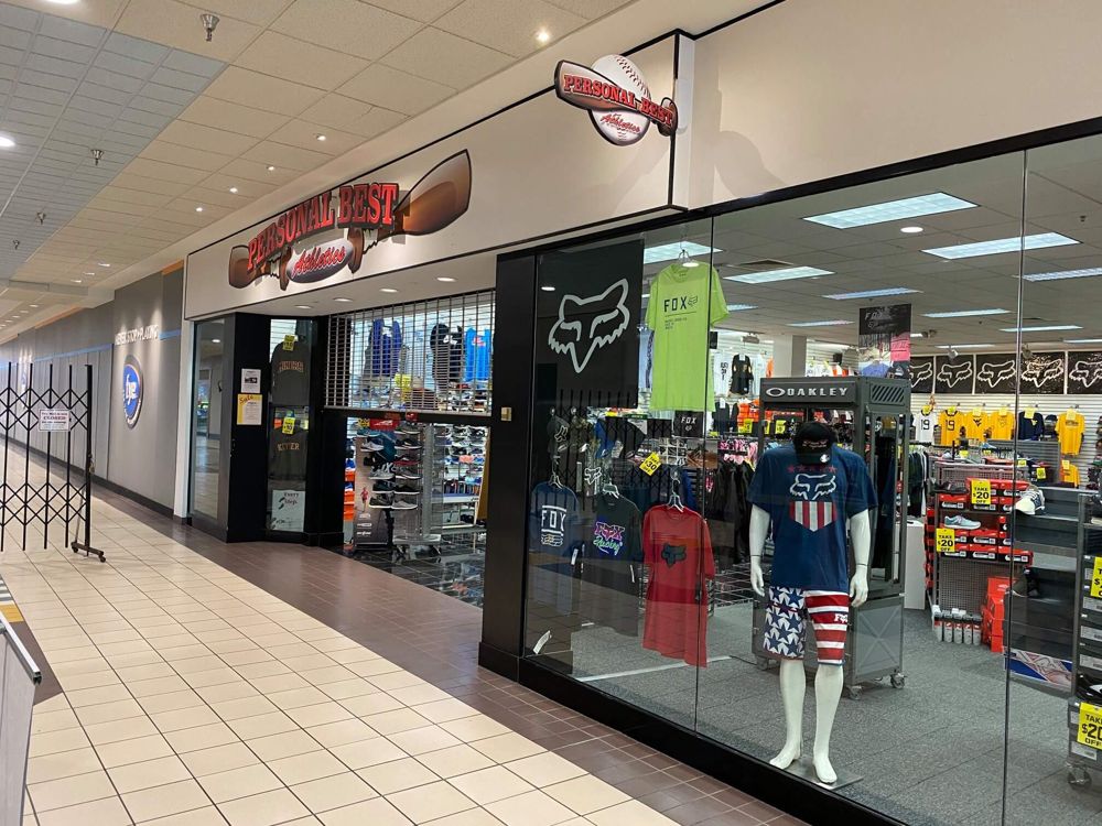 Storefront view of the Personal Best store in the country club mall. The Personal Best signage is in view. The window is full of Fox apparel and behind it other sporting apparel can be seen. Across from the store is a sign for F.Y.E.