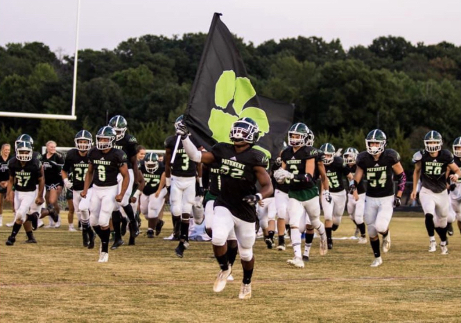 Patuxent high school football team charges across a grass football field together. The player in front leading the charge holds a large black flag with a lime-green paw print on it.
